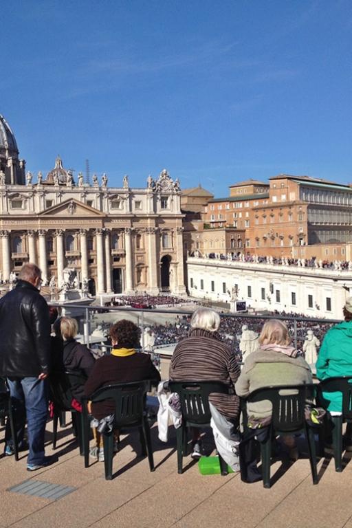 Im Vordergrund eine Terrasse, mehrere Personen sitzen in Gartenstühlen an der Brüstung und schauen auf den Petersplatz. Im Hintergrund der Petersdom