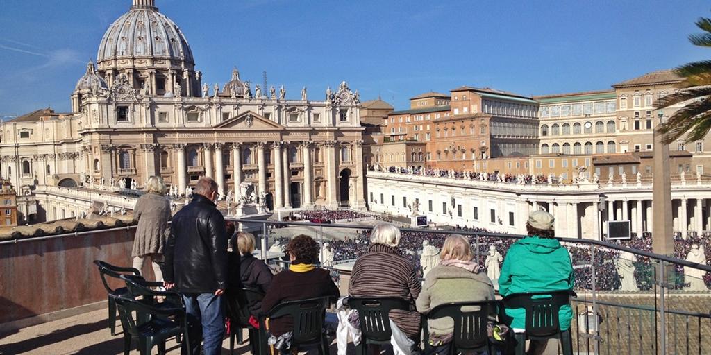 Im Vordergrund eine Terrasse, mehrere Personen sitzen in Gartenstühlen an der Brüstung und schauen auf den Petersplatz. Im Hintergrund der Petersdom