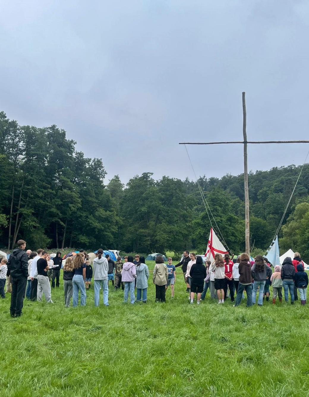 Im Fokus ist ein großes einfaches Kreuz aus zwei Baumstämmen in einem Zeltlager auf einer Wiese