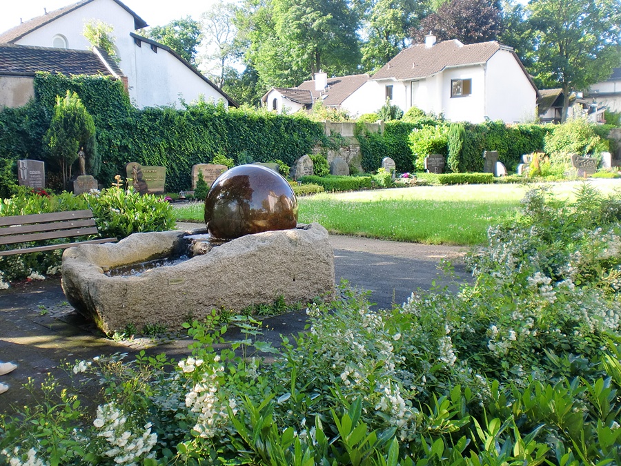 Blick auf einen Brunnen mit Sitzbank, im Hintergrund sind Grabstätten vor der Friedhofsmauer zu sehen