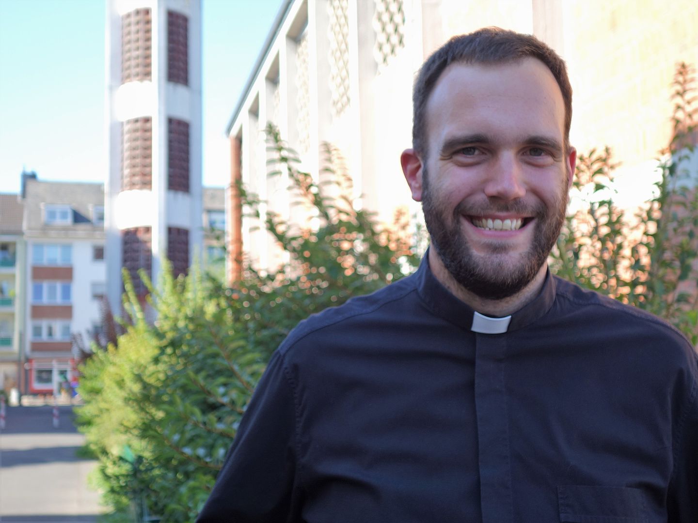 Jugendpfarrer Dominic Lück im schwarzen Hemd mit Priesterkragen im Hintergrund die Kirche St. Elisabeth mit Turm sowie zwei weitere Häuser