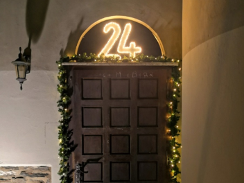 Über einer großen Holztür die Zahl 24, die leuchtet. Die Tür ist mit einer Tannengirlande und Lichterkette geschmückt