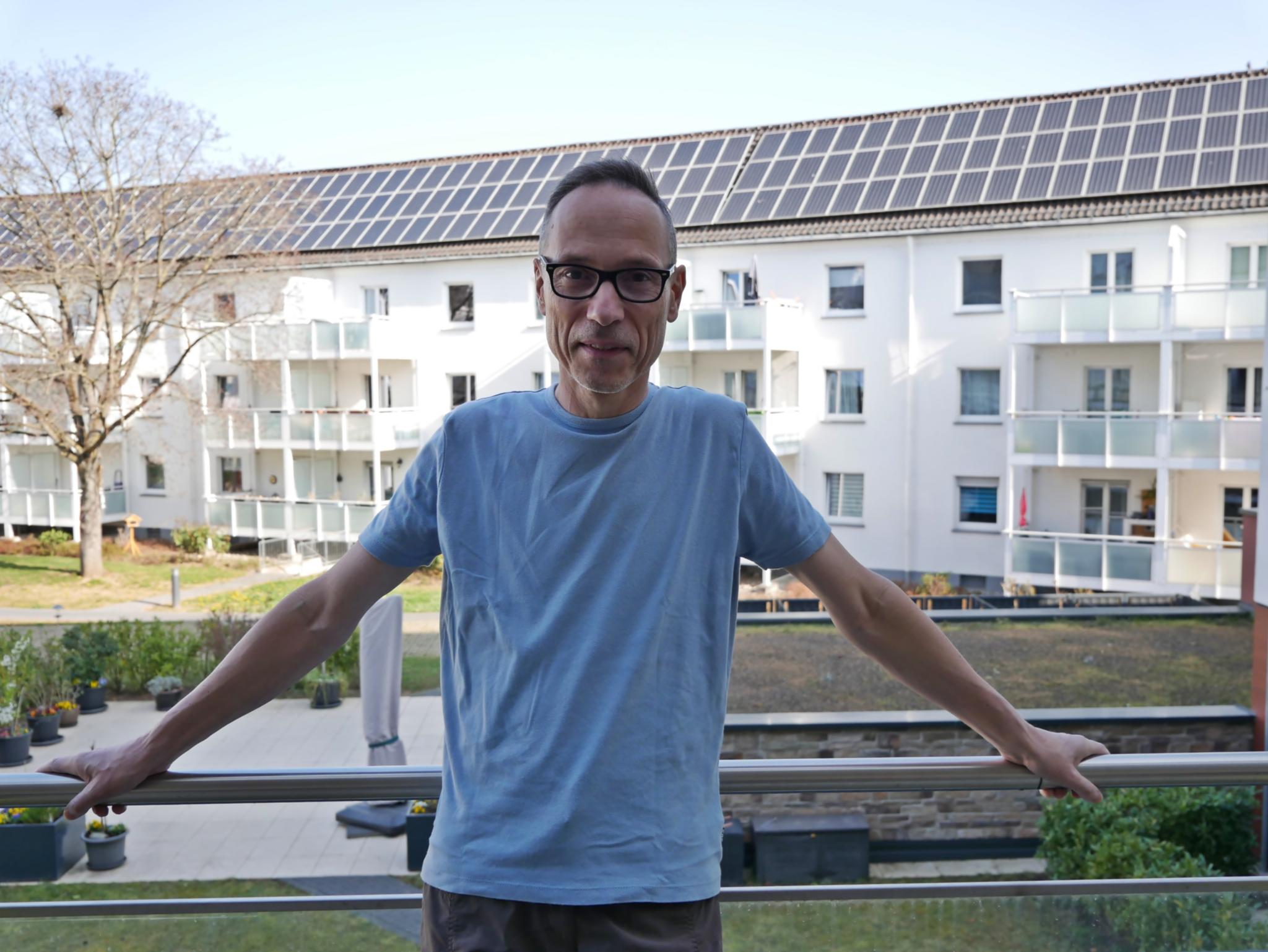 Heinz-Peter Wilbertz ist ab dem Oberkörper zu sehen, er steht auf dem Balkon seiner Wohnung, mit dem Rücken zum Geländer, im Hintergrund der Hof des Mehrgenerationenhauses sowie Mehrfamilienhäuser