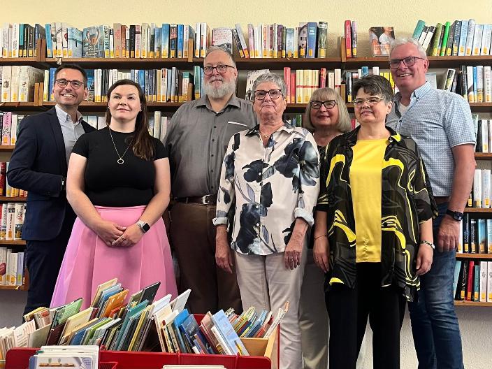 Feierlich gekleidete Personen vor Bücherregalen, davor Kisten mit Büchern