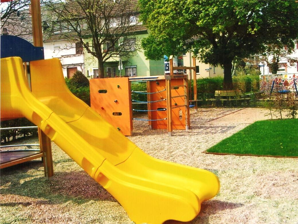 Blick auf das Außengelände der KiTa, mit Rutschen und Sand und einer Bank unter einem Baum