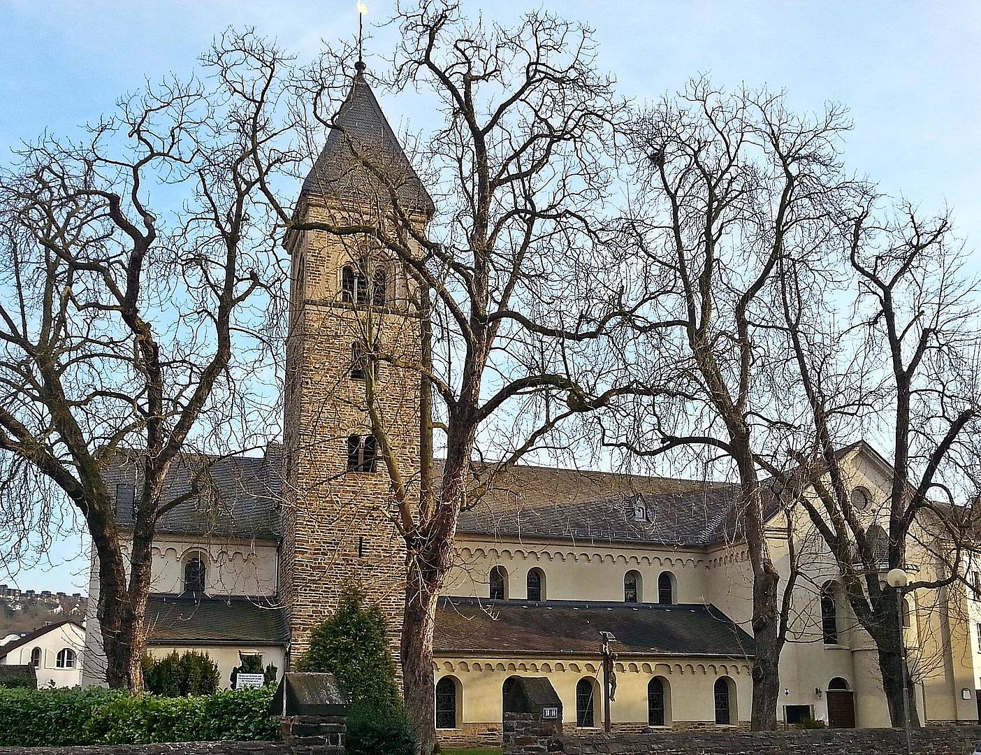 Kirche St. Laurentius von außen, von der Straßenseite her, die großen Kastanien davor haben keine Blätter. Blauer Himmel.