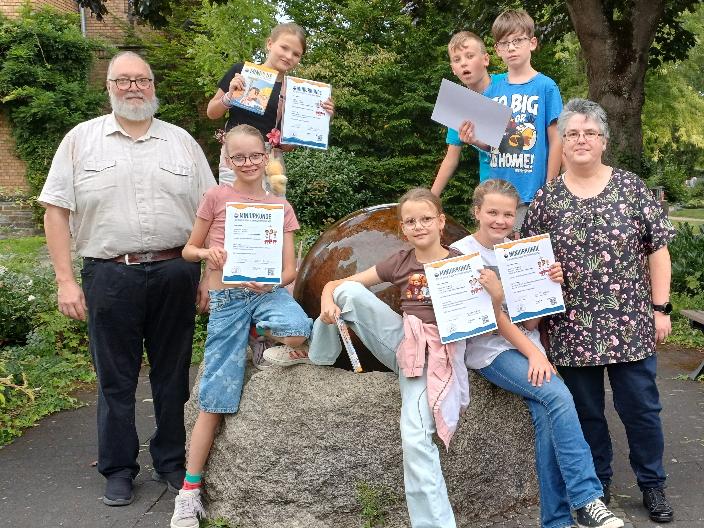 in der Mitte sechs Kinder, die eine Urkunde hochhalten, rechts und links stehen ein Mann und eine Frau