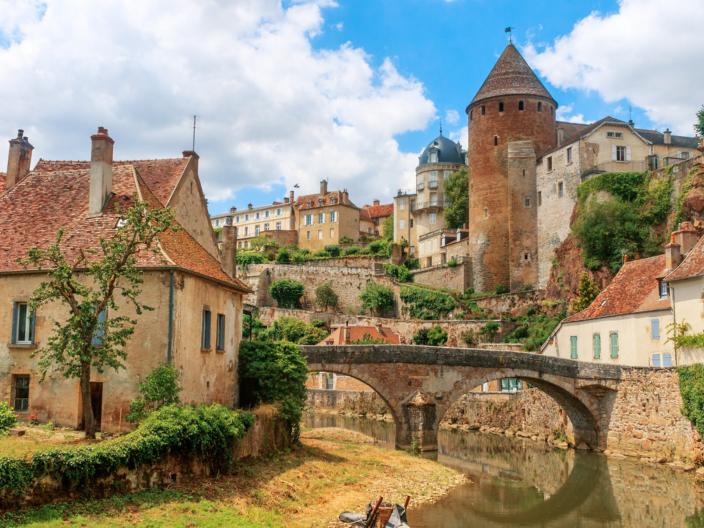 Zu sehen ist eine Ansicht der Stadt Semur, die beidseitig an einem Fluss liegt über den eine mittelalterliche Brücke führt. Idylisch, malerisch sind mittelalterliche Gebäude und prominent ein Turm zu sehen.