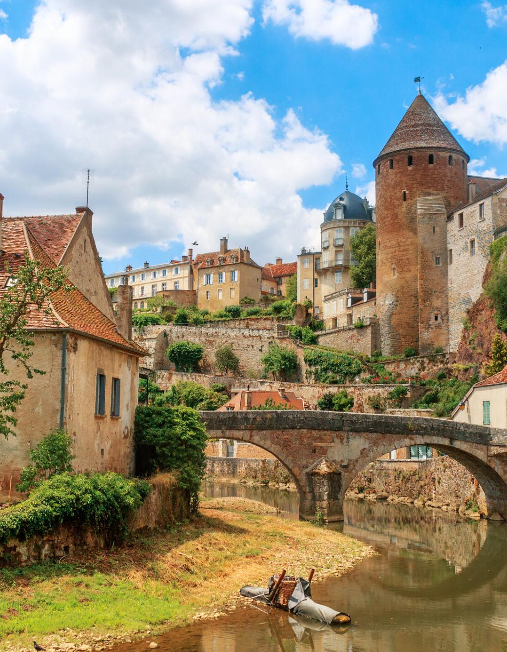 Zu sehen ist eine Ansicht der Stadt Semur, die beidseitig an einem Fluss liegt über den eine mittelalterliche Brücke führt. Idylisch, malerisch sind mittelalterliche Gebäude und prominent ein Turm zu sehen.