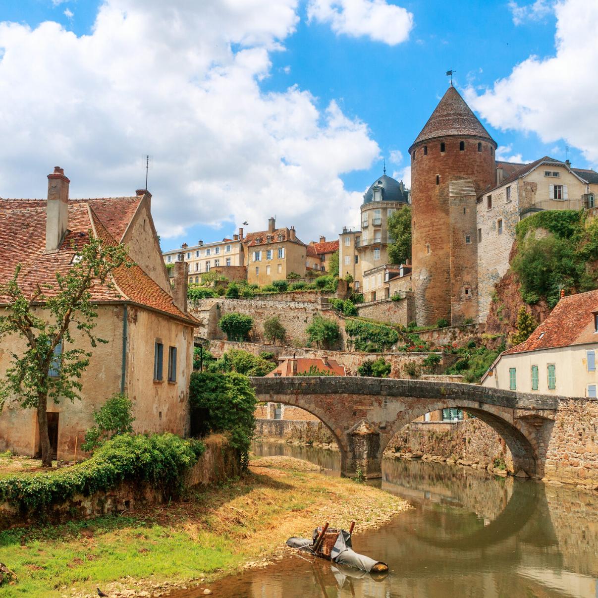Zu sehen ist eine Ansicht der Stadt Semur, die beidseitig an einem Fluss liegt über den eine mittelalterliche Brücke führt. Idylisch, malerisch sind mittelalterliche Gebäude und prominent ein Turm zu sehen.